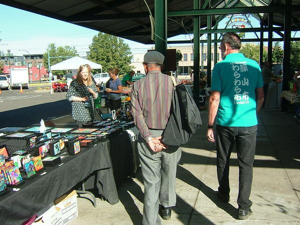 「篠山市わらわらのティシャッ」を着た外国の男性が外のテーブルに置いてある品物を見ている写真